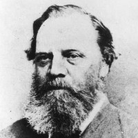 Image: A photographic head-and-shoulders portrait of a middle-aged Caucasian man with a bushy salt-and-pepper beard and receding ear-length hair. He is wearing a late-Victorian suit with bowtie