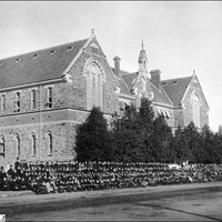 Image: Hundreds of schools students in black uniforms pose in lines outside a large symmetrical stone building with decorative brickwork between floors, two huge arched windows on the front gable ends and smaller windows on the sides.