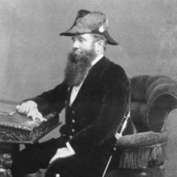 Image: a bearded man in three quarter profile and wearing a hat and sword sits at his writing desk. 