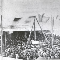 Image: a crowd of people in 1870s clothing gather around a small stage where a large foundation stone is being lowered into place.
