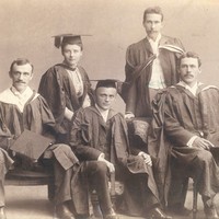 Image: Four men and a woman in university graduation attire sit on chairs and pose for a photograph