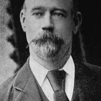 Image: A photographic head-and-shoulders portrait of a balding, middle-aged man with a goatee. The man is wearing an Edwardian-era suit and tie