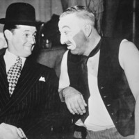 Image:Smiling man in suit and hat next to man in make-up and dirty vest