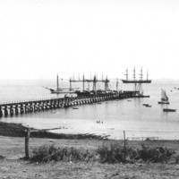 Image: A jetty jutting out into the ocean surrounded by various size sailing ships.