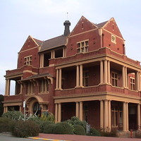 Image: Modern photograph of a large red brick building with yellow painted decorative details such as columns, and featuring a verandah and balconies.