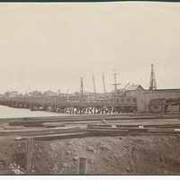 Image: A wooden footbridge extends across a river. A handful of nineteenth-century buildings and sailing ships are visible in the background