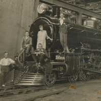 Image: The front of a train sitting in a large shed with five people sitting on the frond looking towards the camera