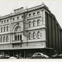 Image: a four storey building with highly decorative facade and plain brick sides. A large arch surrounds a balcony in the centre of the facade and the entrance is protected by a verandah. 1960s era cars are parked outside.