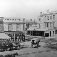 Image:  A three horse team and laden wagon is standing on a dirt street near an intersection. In the centre of the view, a man on a scaffold is engaged in building work. The building on the near side of the intersection has a sign reading "The Bee Hive"