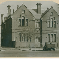 Image: A 1930 era car is parked in front of a plain two storey brick building with gable roofs.