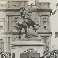 Image: a group of men in dark suits and top hats sit on a stage in front of a statue of a soldier on a horse. A large crowd surrounds them.