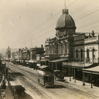 Image: a city street with a number of pedestrians and a horse drawn bus. Towards the centre of the view is an ornate two storey building with a dome. 