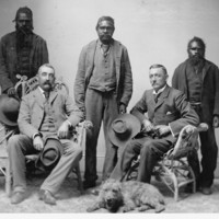 Image: R.T. Maurice’s expedition party. Three Aboriginal men standing, two white men sitting, one dog on the floor. All the men wear suits.