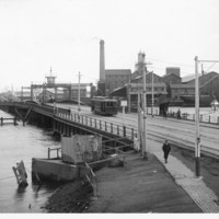 Image: A tram and four pedestrians cross a bridge across a river in a port. A complex of nineteenth-century buildings and a wooden-hulled hulk are visible in the background