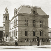 Image: Two women in early 20th century clothing, one with a sun umbrella, stroll past a large stone building with two main floors as well as basement and attic space. 