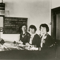 Image: Three women are seated at a switchboard. Behind them is a sign that says "Emergency Calls" and lists numbers for police, fire, ambulance, hospital and waterworks