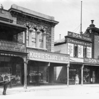 Image: a row of two storey shops with verandahs line a dirt road. To the right a horse drawn cart stands outside a hotel with a balcony.