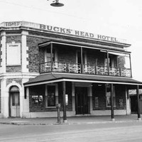 Image: a two storey corner hotel with a small balcony on one side. A verandah protrudes further out to the street beneath the balcony. A parapet sign reads: "TISHERS BUCKS' HEAD HOTEL"