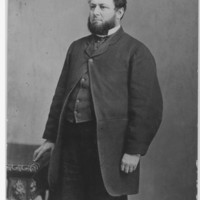 Image: A photographic portrait of a bearded man standing next to a table. The man is wearing an overcoat and other mid-nineteenth century clothing
