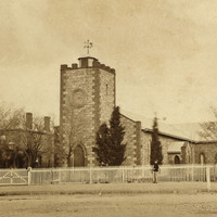 Image: A low, squat church building stands at the intersection of two dirt roads. Two people stand in front of the church