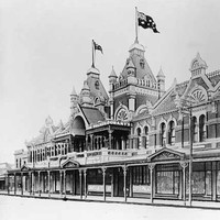 Image: a two storey ornate brick building with arched windows, a series of store windows beneath a verandah, two towers with square cupolas and flagpoles flying Australian flags above the main entrance and a tiled roof