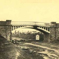 Image: a metal arched bridge with stone abutments passes over a railway track.