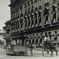 Image: a leisurely scene, with an open horse tram trundling along the road, whilst ladies walk holding sun umbrellas. Two of the ladies are tending to a child in a perambulator. On the right is Eagle Chambers, an imposing three storey building. 