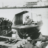 Image: A small motor vessel is launched from its construction slip into a river. A crowd of people in late-Victorian attire look on