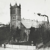 Image: A large stone church building with a tower topped by four spires stands at the corner of two dirt roads. A man stands in front of the churchyard fence