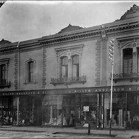 Image: a black and white photograph of a two storey terrace building with large plate glass shop windows on the first floor displaying a range of clothing and fabrics