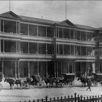 Image: A line of horse drawn vehicles waits outside a huge three storey hotel with balconies on the second and third floors and a verandah on the ground floor. 