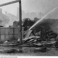 Image: A jet of water is directed at a large, smouldering pile of debris. Burned-out buildings are visible in the background through a haze of smoke