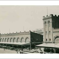 Image: Two large buildings with archway windows and a square-sided tower. Several cars of 1920s vintage are parked in front of the building