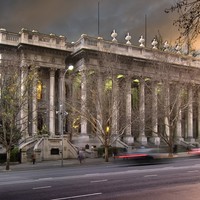 Image: A large stone building with two storey high columns lining its front is lit up by green lights at dusk.