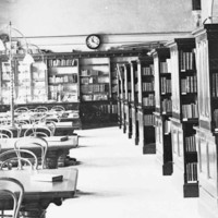 Image: A long, well-lit room containing several shelves of books on one side and six reading tables on the other