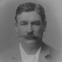 Image: A photographic head-and-shoulders portrait of a man in suit and tie sporting a large handlebar moustache. The frame surrounding the portrait identifies the man as Richard Chaffey Baker