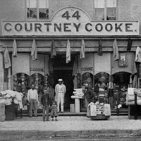 Image: A group of men and children stand outside a two storey terrace building the ground floor of which is serving as a drapery store with fabric sitting in baskets on the street, coats hanging along the storefront and hats in the window. 