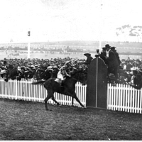 Image: Black and white photo of horseracing in progress, crowd behind a fence