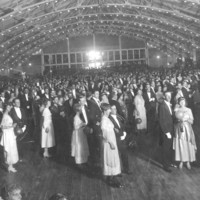 Image: a crowd of men and women wearing 1920s era clothing stand in pairs in a large hall the ceiling of which is decorated with lines of small lights. 