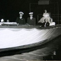 Image: a woman in an extravagant gown sits on a raised dias at the stern of a small boat. Other well dressed individuals sit along the sides of the boat. 