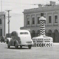 Image: Cars of 1950s vintage pass around a large plinth in the centre of an intersection in an urban area. The words ‘Keep Left’ are painted on the plinth, as are several large left-facing black arrows 