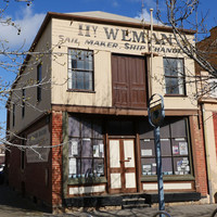 Image: A two-storey building with brick first floor and corrugated metal second floor. The words ‘Hy. Weman, Sail Maker, Ship Chandler’ are painted on the building shop front