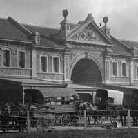 Image: A long two-storey brick terrace building with three large double height arches and decorative gables. Along the street at the front a number of horse drawn carts are parked. 