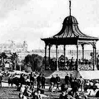 Image: A crowd of people in early twentieth century attire sit on the grass around a rotunda where a band plays