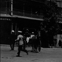 Image: a series of three storey buildings line one side of a wide city street. Between the buildings and the road is a line of trees. Travelling along the street are horse-drawn vehicles, motor cars and an electric tram, as well as people in 1920s clothes