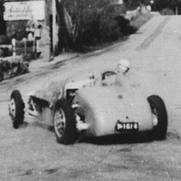 Image: A 1950s vintage racing car drives around a corner and through the streets of a small rural town. A handful of small historic stone buildings are visible on either side of the road