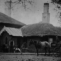 Image: man stands with dog in front of a brick house with a thatched roof. Beside him sits a woman while another man drives a horse drawn buggy.