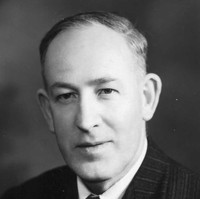 Image: A photographic head-and-shoulders portrait of a clean-shaven, middle-aged Caucasian man wearing a suit of 1940s vintage