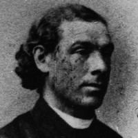 Image: A photographic head-and-shoulders portrait of a young Caucasian man in a Victorian-era Catholic clergyman’s outfit. The man has long, dark wavy hair pulled back behind his head