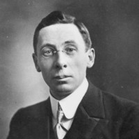 Image: A photographic head-and-shoulders portrait of a young man wearing a suit and wire-rimmed spectacles